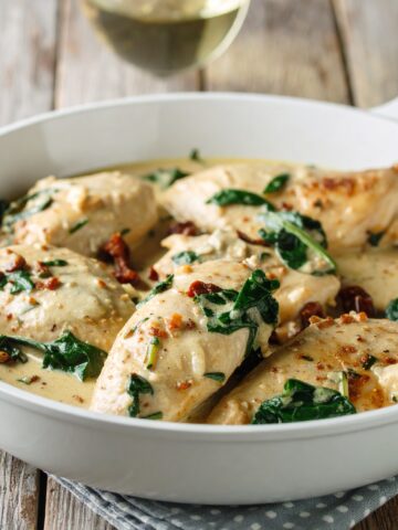 A large white skillet filled with golden chicken breasts in a creamy sauce, flecked with sun-dried tomatoes and spinach. Served on a rustic wood table with a glass of white wine nearby.